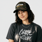 Camo Mesh Trucker Cap worn by woman with shoulder-length dark hair, featuring white 'HORIZN' logo and gold 'MotoCo' graphic, styled with gray 'CHASE THE HORIZN' t-shirt.